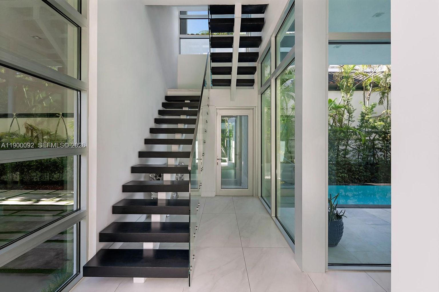This modern interior space features a striking floating staircase with dark treads and a glass railing, serving as the focal point of the hallway. Large, floor-to-ceiling windows on both sides of the corridor flood the area with natural light and provide a clear view of an outdoor swimming pool and lush greenery. The minimalist design is complemented by light-colored, large-format tile flooring and a clean, white aesthetic that emphasizes the home's architectural openness.