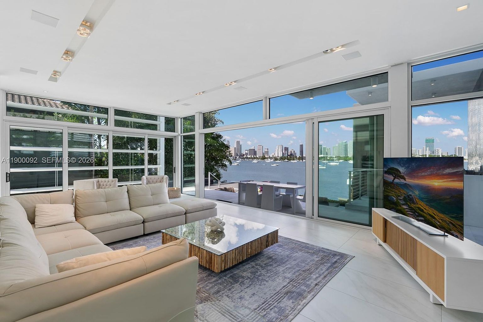 This bright and airy living room features a large, light-colored sectional sofa arranged around a glass-topped coffee table with a textured base. The space is defined by expansive floor-to-ceiling windows that provide an unobstructed, panoramic view of the bay and city skyline. A modern media console with light wood accents sits below a large television, while the room's minimalist aesthetic is complemented by polished light-toned flooring and a neutral area rug.