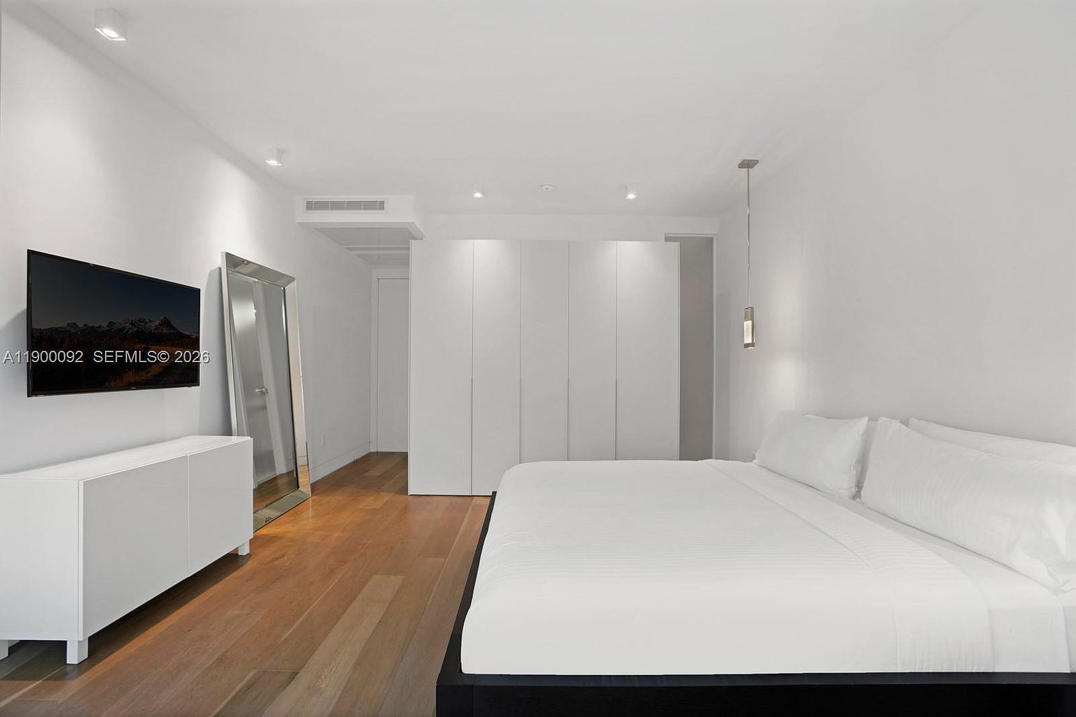 This minimalist primary bedroom features a clean, monochromatic white aesthetic with warm wood flooring. The room is furnished with a large bed, a sleek white media console topped with a wall-mounted television, and a tall, framed floor mirror. A wall of seamless white cabinetry provides ample storage, while recessed lighting and a single pendant light create a bright and airy atmosphere.