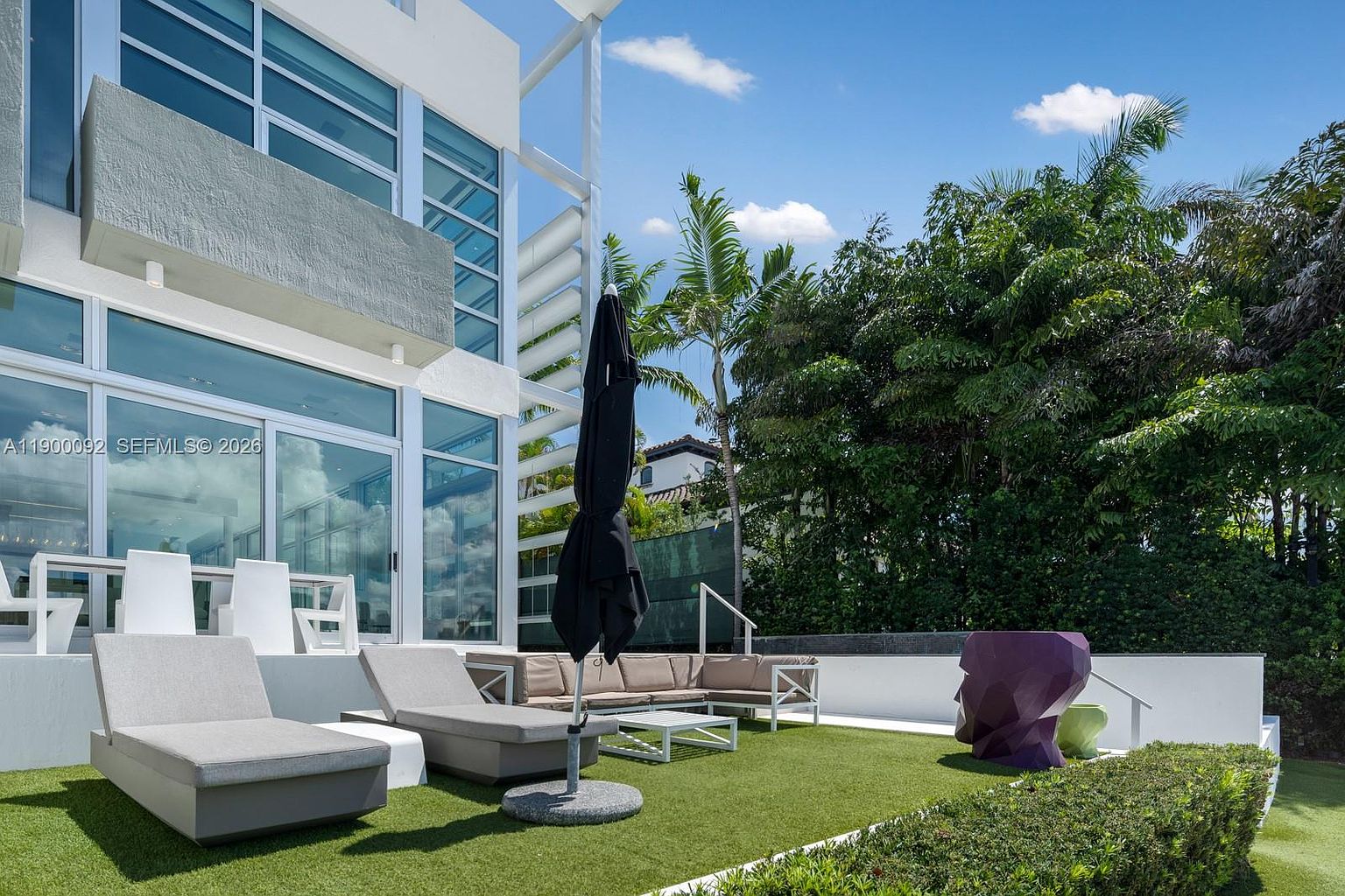 This modern outdoor living space features a synthetic turf lawn furnished with two lounge chairs, a sectional sofa, and a central umbrella. The area is adjacent to a contemporary home with large glass panels that provide a seamless transition between indoor and outdoor living. A unique, abstract purple sculpture adds a bold artistic element to the landscaped garden setting.