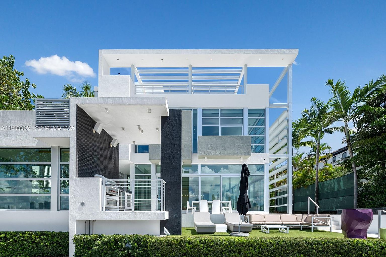This modern, multi-level residence features a striking architectural design with a mix of white stucco and dark textured accents. The rear exterior showcases large glass sliding doors that open onto a manicured lawn, complete with outdoor lounge furniture and a black patio umbrella. A prominent white pergola structure tops the third level, adding a geometric flair to the contemporary aesthetic.