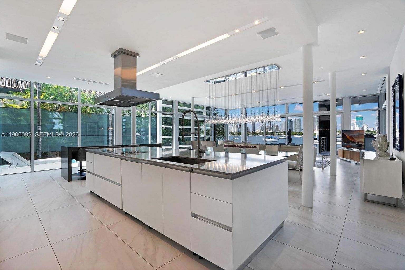 This expansive, modern kitchen features a large central island with a stone countertop and integrated sink, serving as the focal point of an open-concept living area. The space is defined by sleek white cabinetry, a stainless steel range hood, and floor-to-ceiling windows that provide panoramic views of the waterfront and city skyline. A dramatic, multi-pendant light fixture hangs above the dining area, enhancing the bright, airy atmosphere created by the neutral color palette and polished tile flooring.