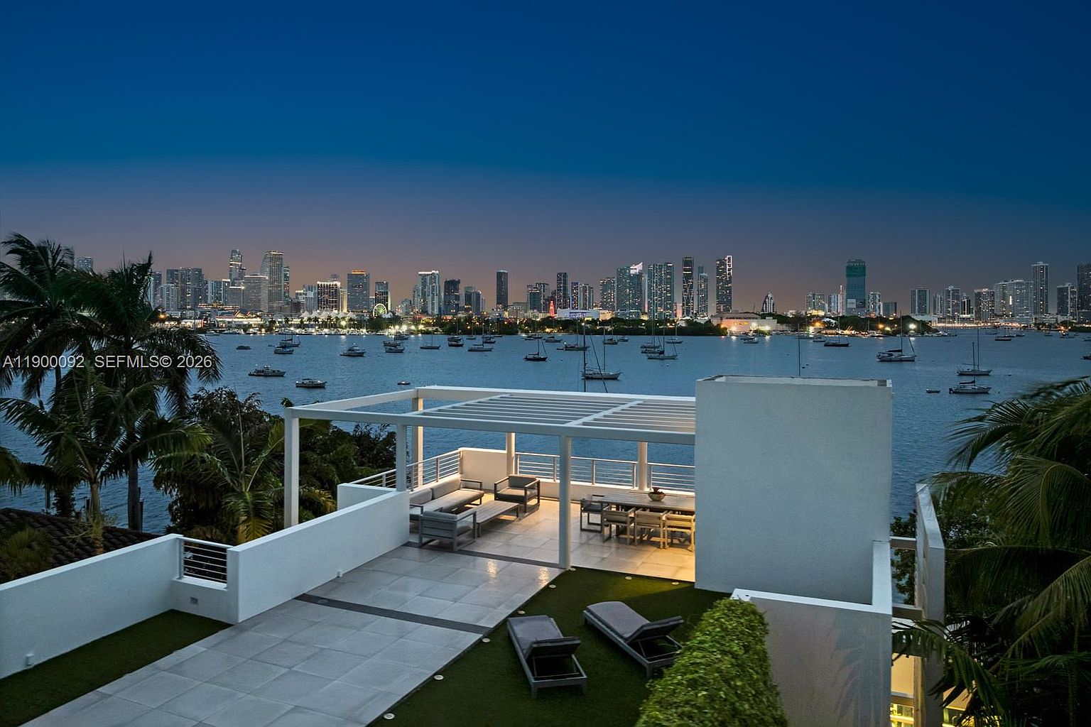 This elevated rooftop terrace offers a sophisticated outdoor living space with a pergola-covered lounge area and a separate dining section. The deck features a mix of light-toned stone tiling and artificial turf, complemented by two lounge chairs positioned for optimal views. The scene is set against a stunning twilight backdrop of a city skyline across the water, creating a luxurious and serene atmosphere.