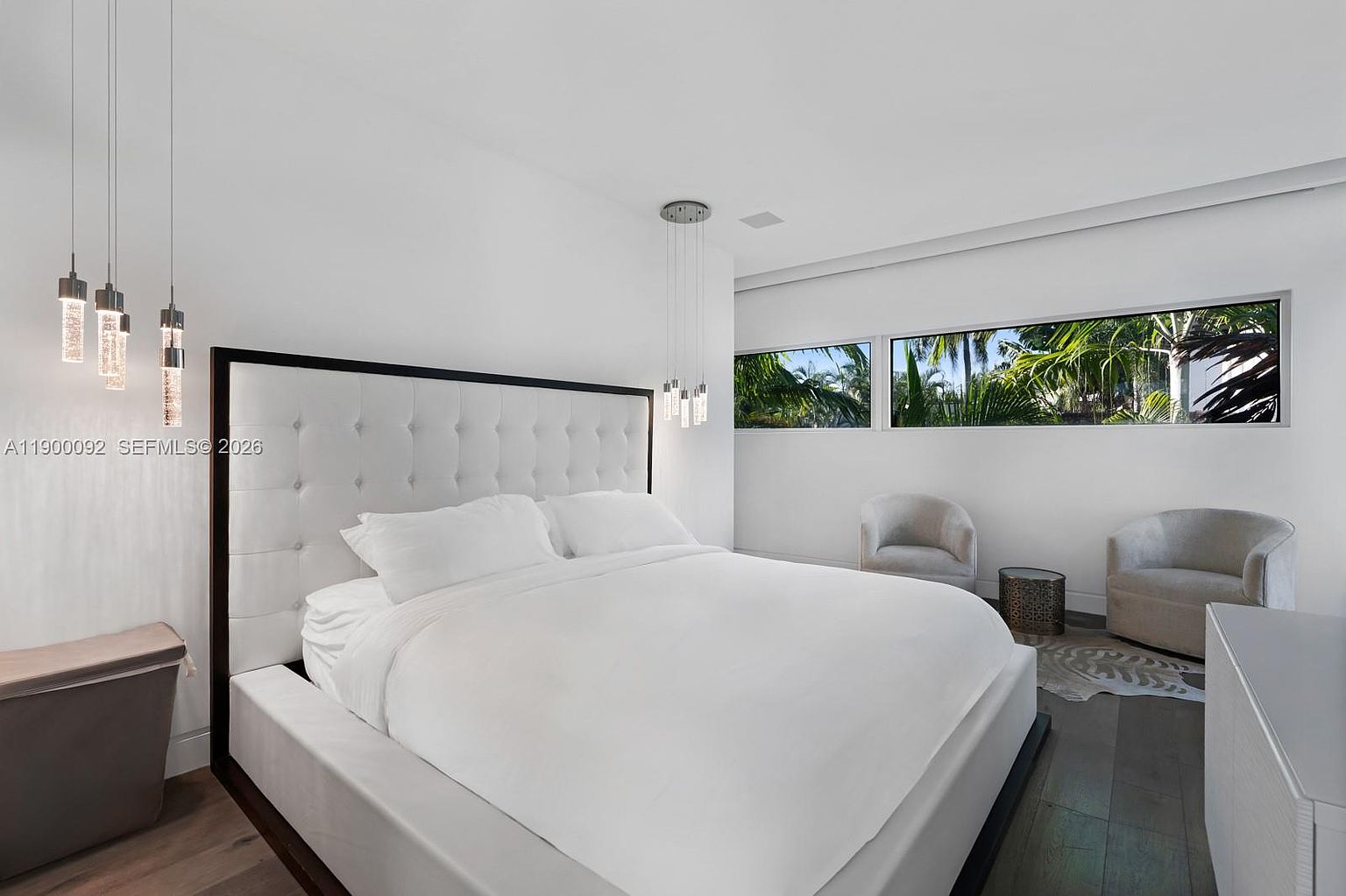 This modern primary bedroom features a large, tufted white headboard as the focal point, complemented by minimalist white bedding and dark wood flooring. The room is illuminated by contemporary pendant lights and natural light streaming through a horizontal row of windows that offer a view of lush greenery outside. Two soft-toned armchairs and a small side table create a cozy seating area, contributing to the serene and sophisticated atmosphere of the space.