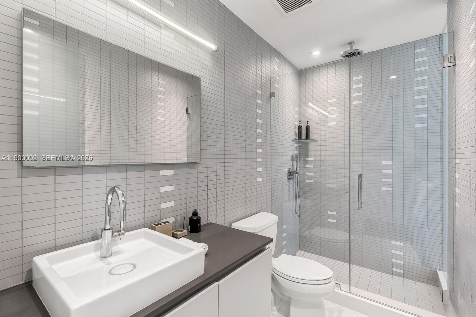 This modern, minimalist bathroom features a sleek vanity with a rectangular sink and a dark countertop, complemented by a large, frameless mirror. The walls are finished with light gray, narrow rectangular tiles that extend into the walk-in shower, which is enclosed by a clear glass panel. A rainfall showerhead and a handheld sprayer are mounted within the shower, creating a clean and contemporary aesthetic.