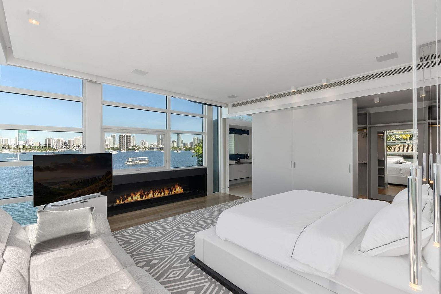 This modern primary bedroom features a minimalist aesthetic with a large white bed and a patterned area rug. A linear fireplace is built into the wall beneath expansive windows that offer a panoramic view of the water and city skyline. The room includes a sleek seating area and leads into a walk-in closet, creating a bright and airy atmosphere.