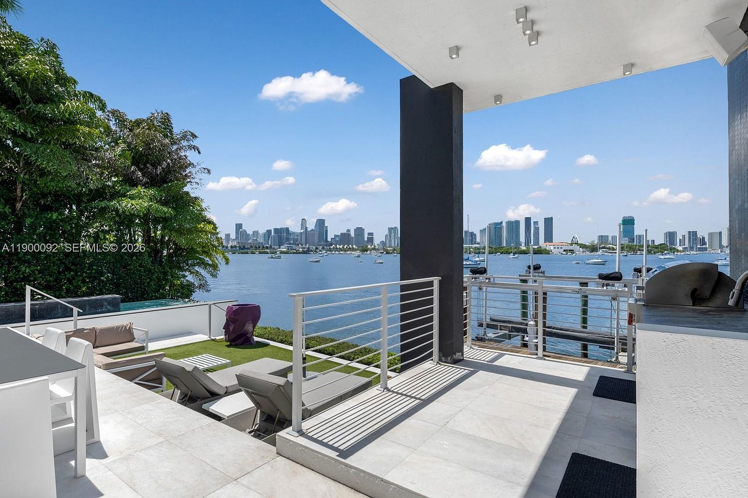 This luxurious waterfront terrace offers a panoramic view of a city skyline across the bay, featuring a sleek, modern design with light-toned stone flooring. The space includes a covered lounge area with a grill, a lower deck with sun loungers, and a small infinity-edge pool overlooking the water. The perspective is from an elevated vantage point, capturing the seamless transition between the private outdoor living space and the expansive coastal scenery.