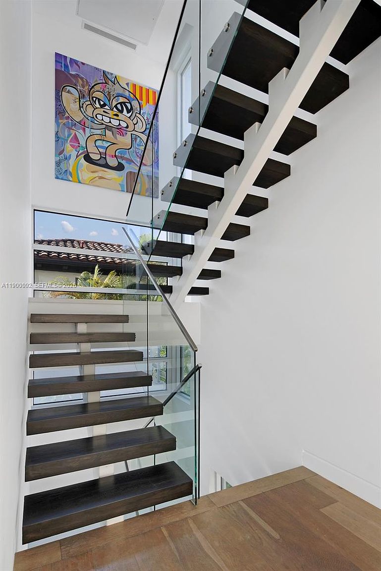 This modern staircase features dark wood treads and a sleek glass railing, creating an open and airy aesthetic. A vibrant, colorful pop-art style painting hangs on the white wall, adding a bold artistic focal point to the landing. The perspective looks up the staircase, highlighting the clean lines and the natural light streaming in from the window at the landing.