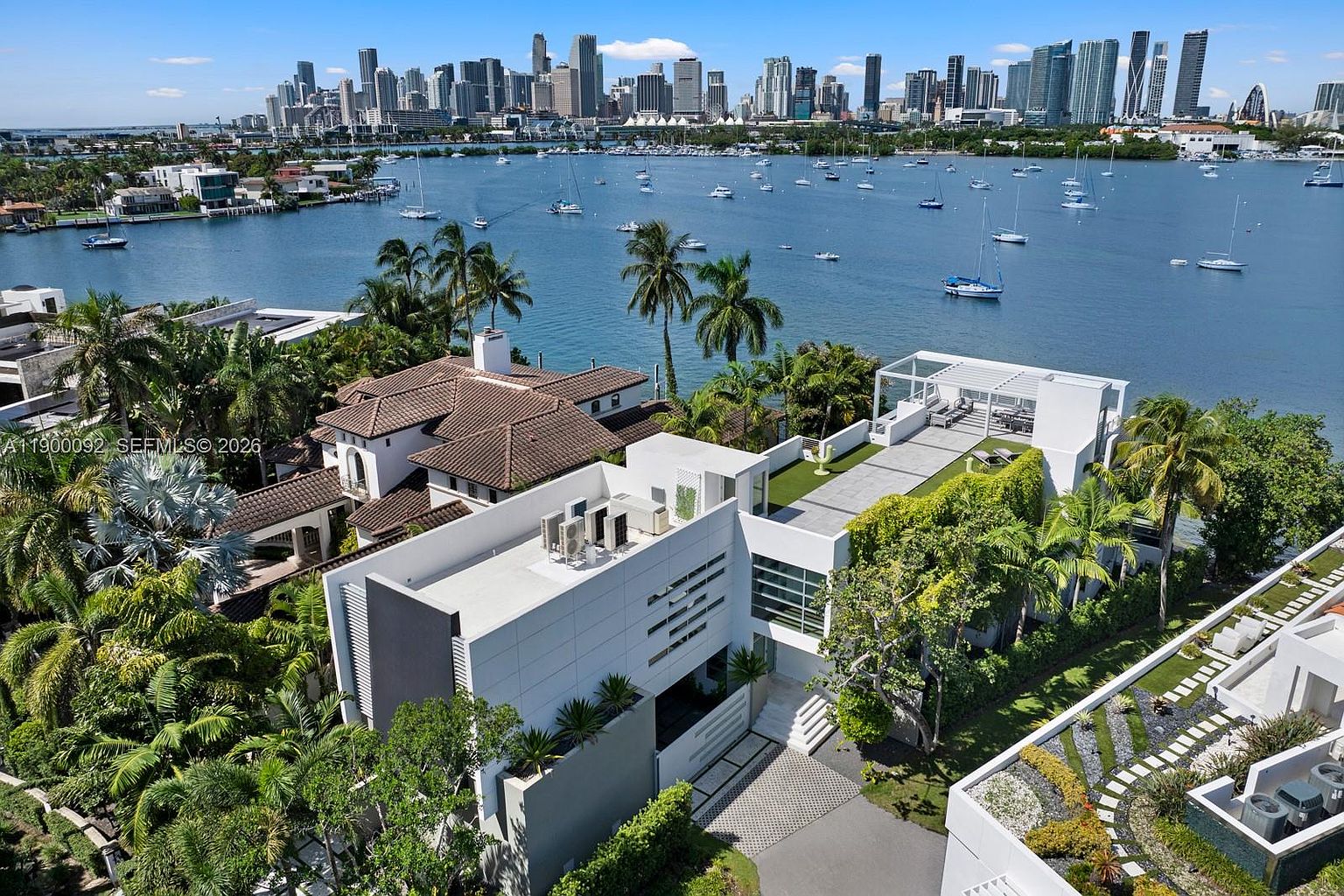 This high-angle aerial shot captures a luxurious waterfront estate featuring a modern, multi-level white residence with a flat roof and a rooftop terrace. The property is nestled among lush tropical landscaping and sits directly on the water, offering a panoramic view of the distant city skyline across the bay. The scene highlights the seamless integration of contemporary architecture with its scenic coastal environment.