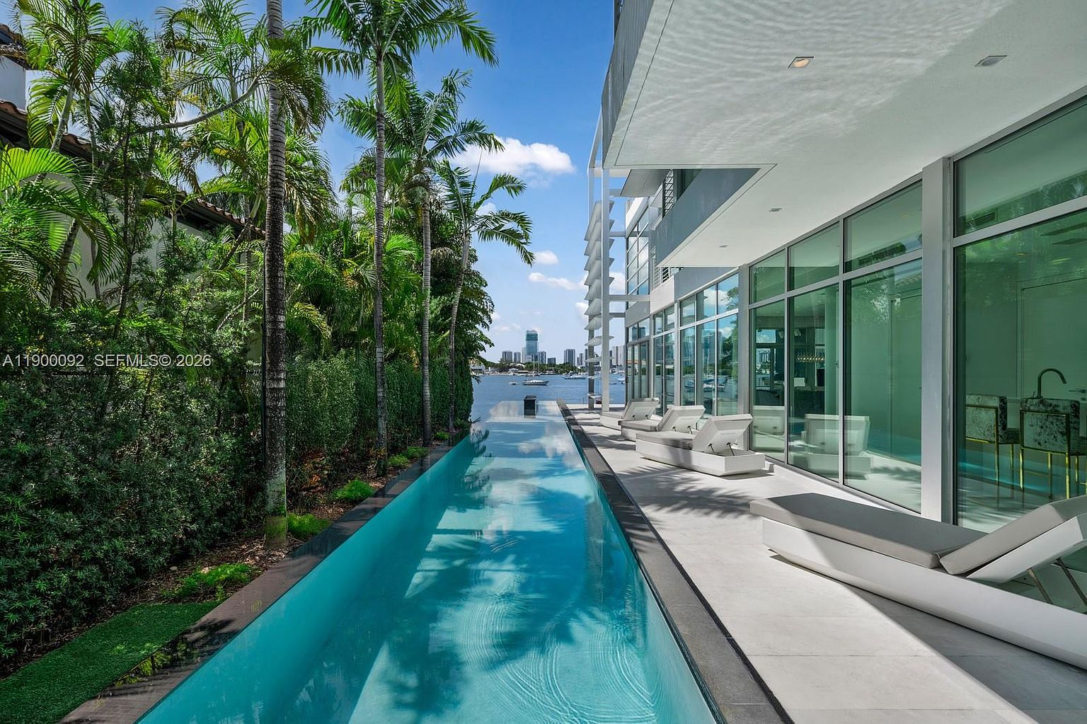 This image captures a sleek, modern lap pool stretching alongside a contemporary residence with expansive glass walls. The pool deck features minimalist lounge chairs, while lush palm trees and dense greenery line the property edge, framing a scenic view of the distant city skyline across the water. The perspective is low and wide, emphasizing the seamless transition between the indoor living space and the outdoor waterfront environment.