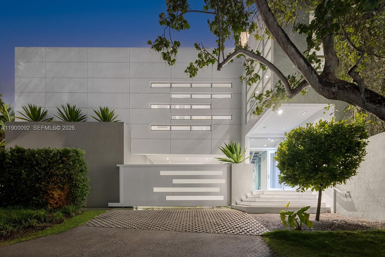 This modern, minimalist residence features a striking facade with clean, geometric lines and a neutral color palette. The front entrance is accessed via a set of wide, illuminated steps, complemented by a gated driveway and lush, manicured landscaping. The architectural design emphasizes symmetry and contemporary aesthetics, creating a sophisticated and welcoming curb appeal.