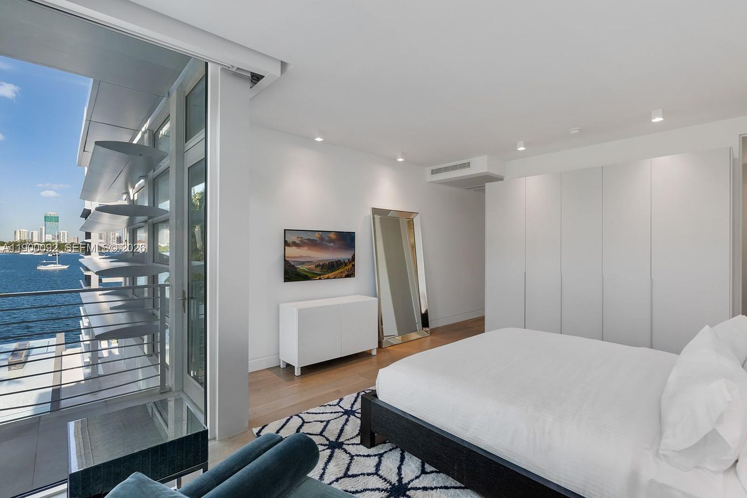 This modern primary bedroom features a minimalist aesthetic with crisp white walls, light wood flooring, and a large bed with a dark frame. A wall of floor-to-ceiling glass doors opens onto a private balcony overlooking the water, while a sleek white console and a tall, leaning floor mirror add functional elegance. The room is completed by a wall of integrated white cabinetry, creating a clean, seamless look that emphasizes the bright, airy atmosphere.