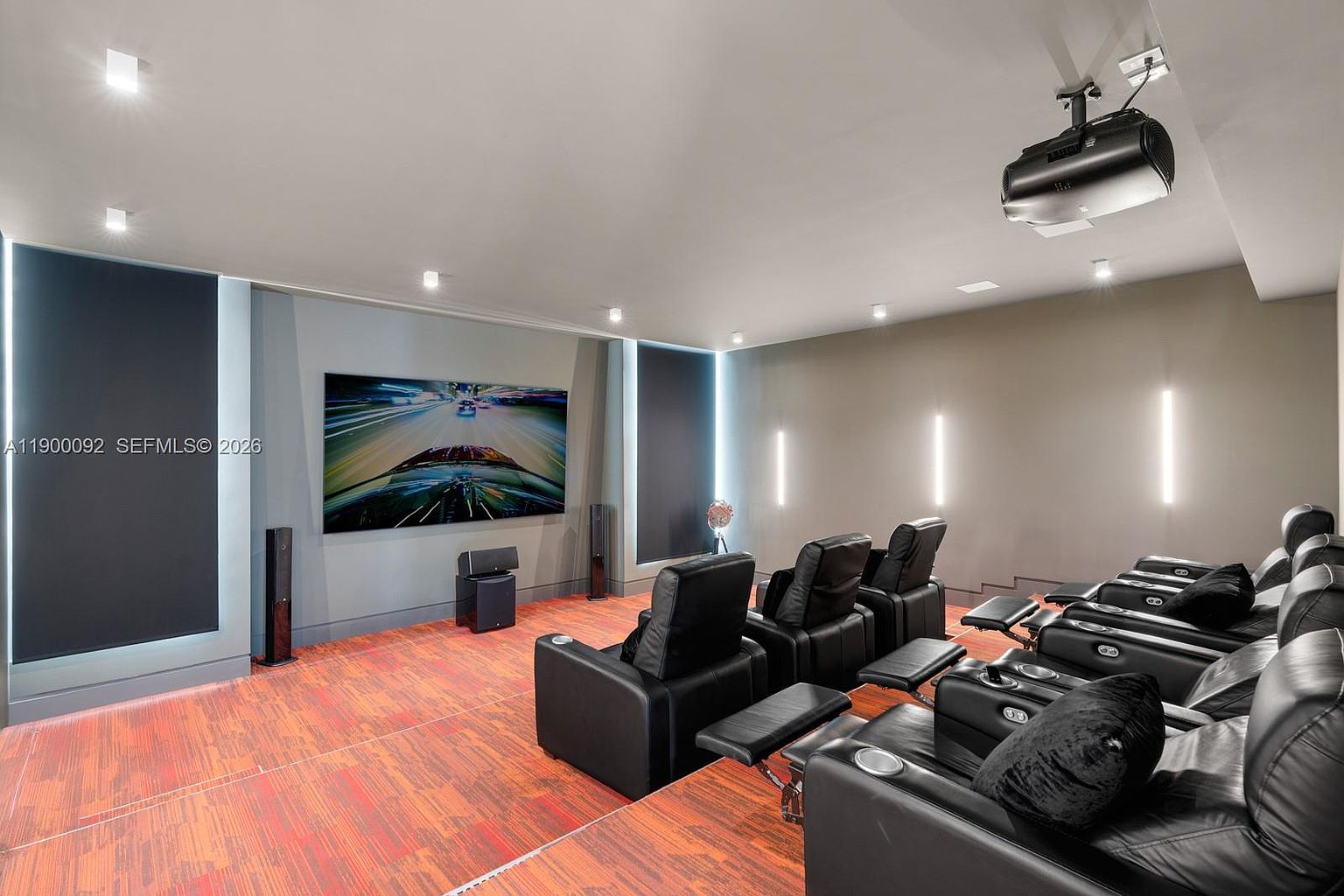 This dedicated home theater room features tiered seating with multiple rows of black leather recliners facing a large wall-mounted television. The space is designed for cinematic immersion, with dark acoustic wall panels, recessed lighting, and a projector mounted on the ceiling. The room is finished with a vibrant, patterned carpet that adds a bold visual element to the otherwise sleek, modern entertainment environment.