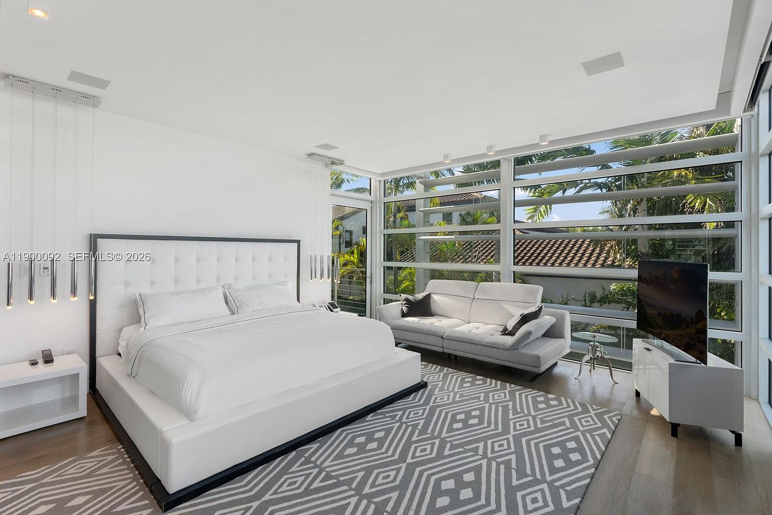 This modern primary bedroom features a large, tufted white headboard as the focal point, complemented by minimalist bedside tables and hanging pendant lights. A light gray sofa sits atop a geometric patterned rug, positioned near expansive windows that offer a view of the surrounding greenery. The room is bright and airy, with light wood flooring and a clean, contemporary aesthetic.