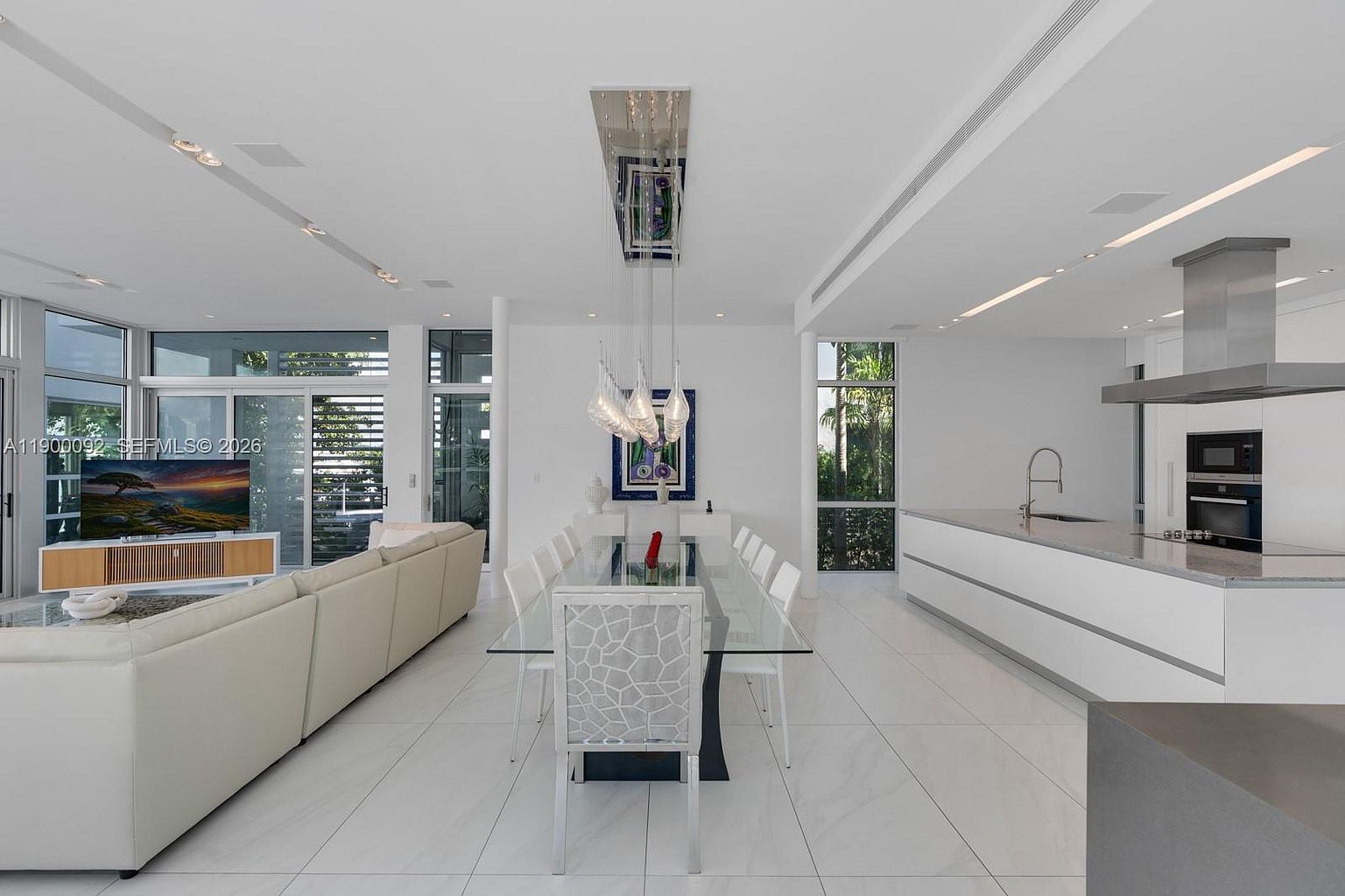 This open-concept living and dining area features a bright, minimalist aesthetic with white walls and large, light-colored floor tiles. A glass-topped dining table with patterned chairs sits beneath a modern, multi-pendant light fixture, while a spacious white kitchen island with a sink and a nearby sofa area complete the expansive, airy layout. The space is illuminated by natural light streaming through large windows that offer views of the surrounding greenery.