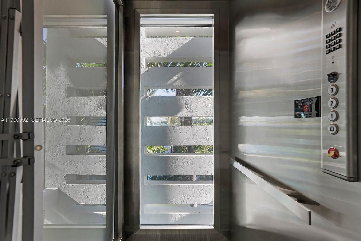 This image shows the interior of a residential elevator, featuring a sleek stainless steel control panel with numbered buttons and a prominent emergency stop switch. A narrow, vertical window with horizontal slats provides a view of the exterior landscape, creating a play of light and shadow within the metallic space. The perspective is from inside the elevator looking out, emphasizing the modern, industrial aesthetic of the lift's design.
