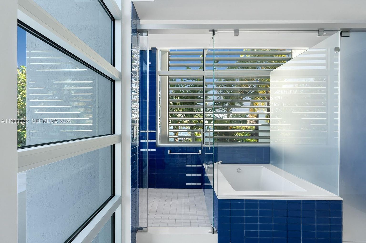 This modern bathroom features a deep soaking tub set against a vibrant blue tiled wall, creating a striking visual contrast. A glass-enclosed shower area is adjacent to the tub, while large windows with horizontal louvers allow for natural light and privacy. The space is characterized by its clean lines, minimalist aesthetic, and a bright, airy atmosphere.
