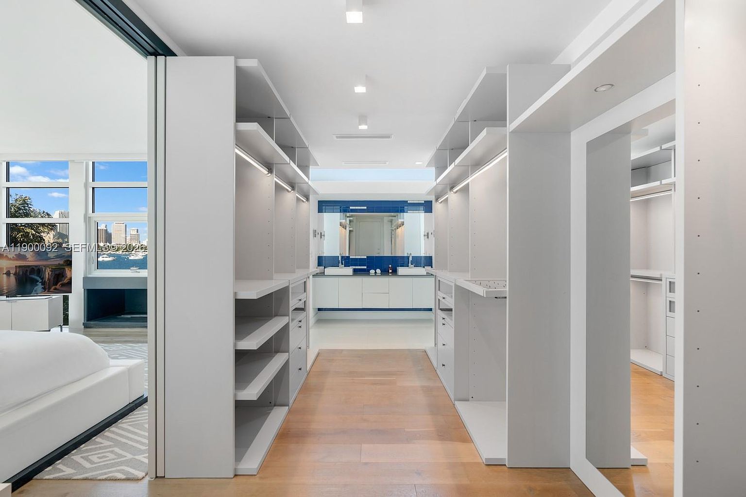 This spacious, modern walk-in closet features custom white cabinetry with integrated lighting and open shelving, leading directly into a bright, blue-accented bathroom. The room is finished with warm-toned wood flooring and a clean, minimalist aesthetic. The perspective looks straight through the closet corridor, framing the vanity area in the background.