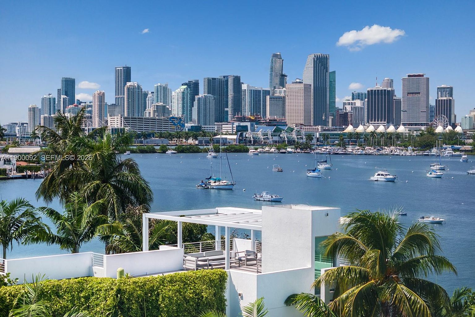 This elevated perspective captures a modern rooftop terrace featuring a white pergola structure and outdoor seating, overlooking a serene bay filled with sailboats. In the background, a prominent city skyline rises against a clear blue sky, creating a striking contrast between the tranquil waterfront and the urban environment. The composition emphasizes the property's prime location and expansive, scenic views.
