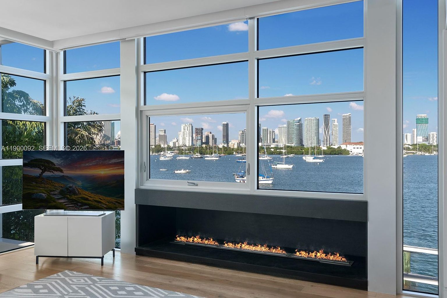 This bright living room features expansive windows that frame a panoramic view of the bay and city skyline. A sleek, linear fireplace is set into a dark, minimalist base, providing a modern focal point beneath the glass. The room is finished with light wood flooring and a patterned area rug, creating a sophisticated and airy atmosphere.
