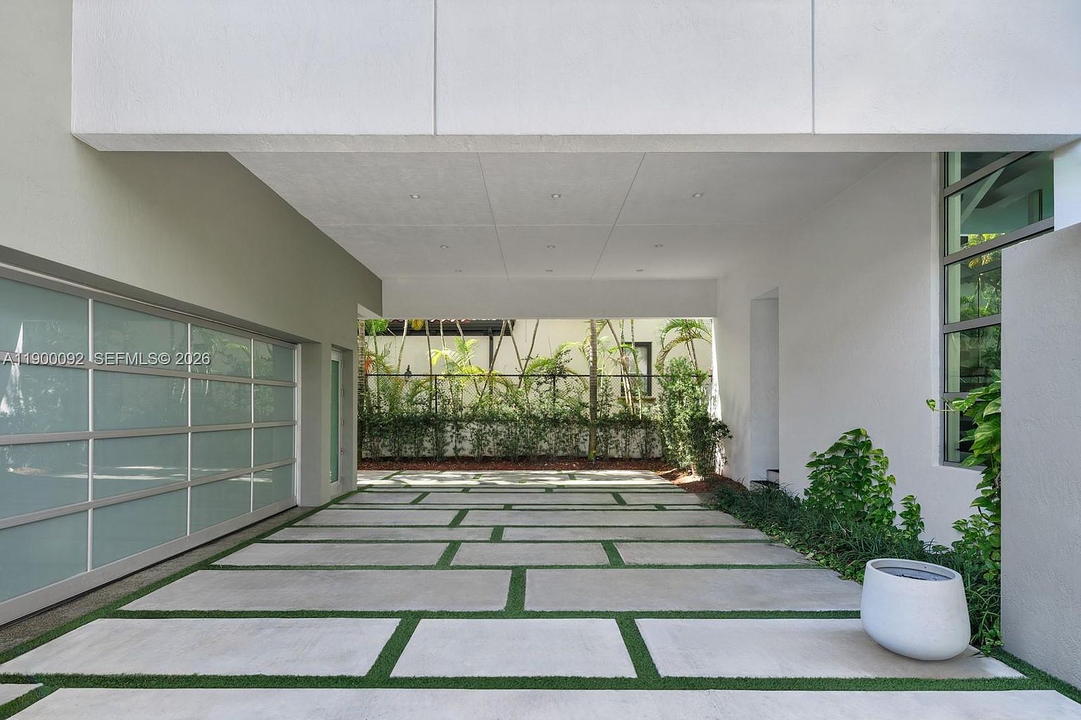 This modern carport area features a sleek, multi-panel glass garage door and a clean, minimalist aesthetic. The ground is paved with large concrete pavers separated by strips of green grass, leading toward a landscaped hedge and fence in the background. The space is covered by a wide, white overhang, creating a bright and sheltered transition area.