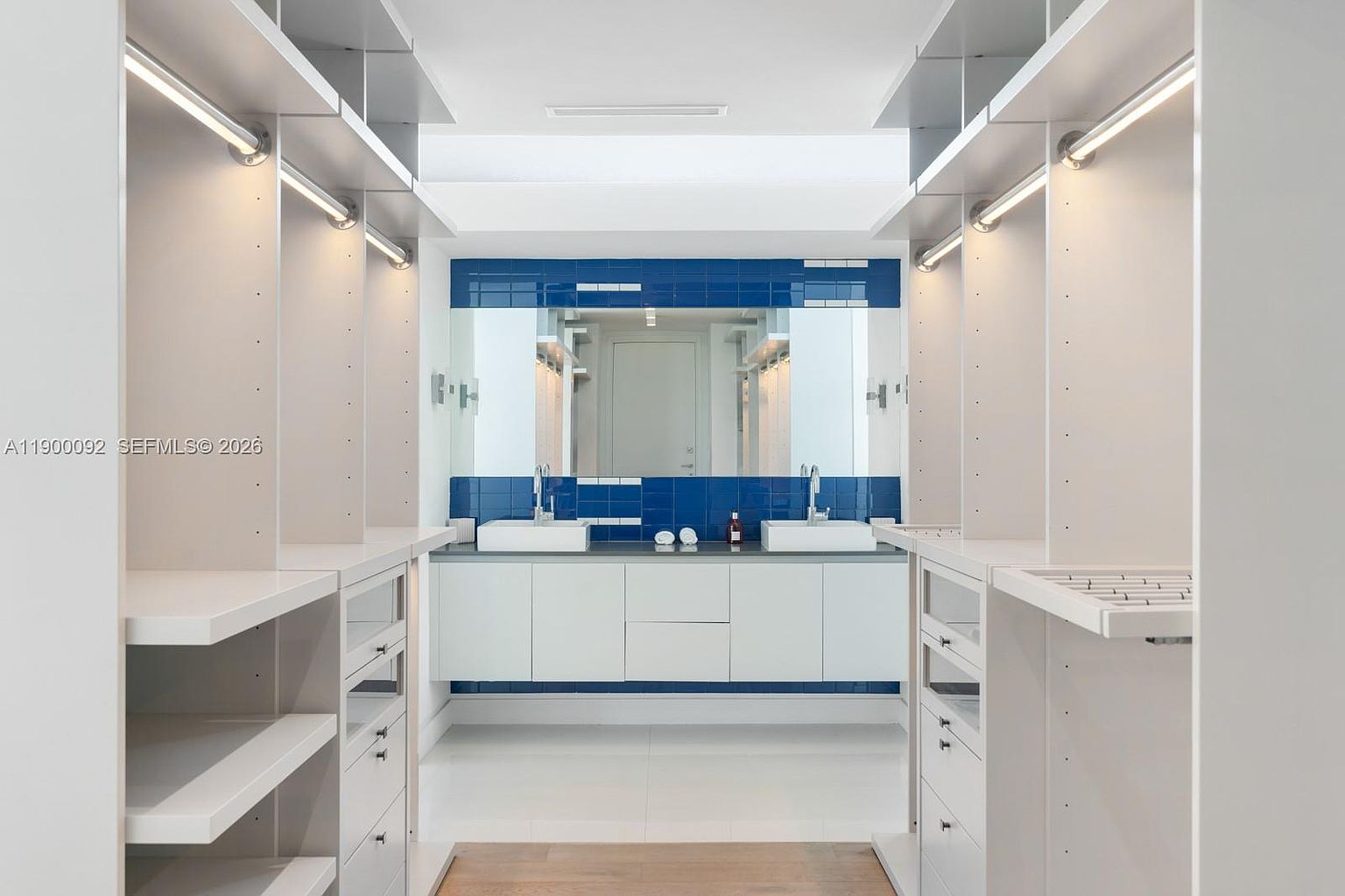 This unique space functions as a hybrid walk-in closet and bathroom, featuring extensive white cabinetry and shelving lining both sides of the room. At the far end, a double vanity with two sinks is set against a striking blue tiled wall and a large mirror. The room is illuminated by integrated lighting strips along the shelving units, creating a bright and modern aesthetic.