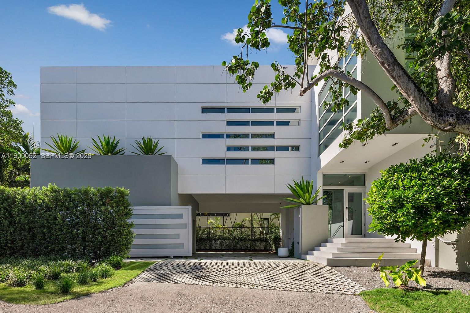 This modern architectural residence features a striking white facade with horizontal window accents and a prominent covered driveway area. The front entrance is elevated by a set of concrete steps, complemented by lush landscaping and a paved driveway. The composition captures a clean, contemporary aesthetic with a focus on geometric lines and natural light.