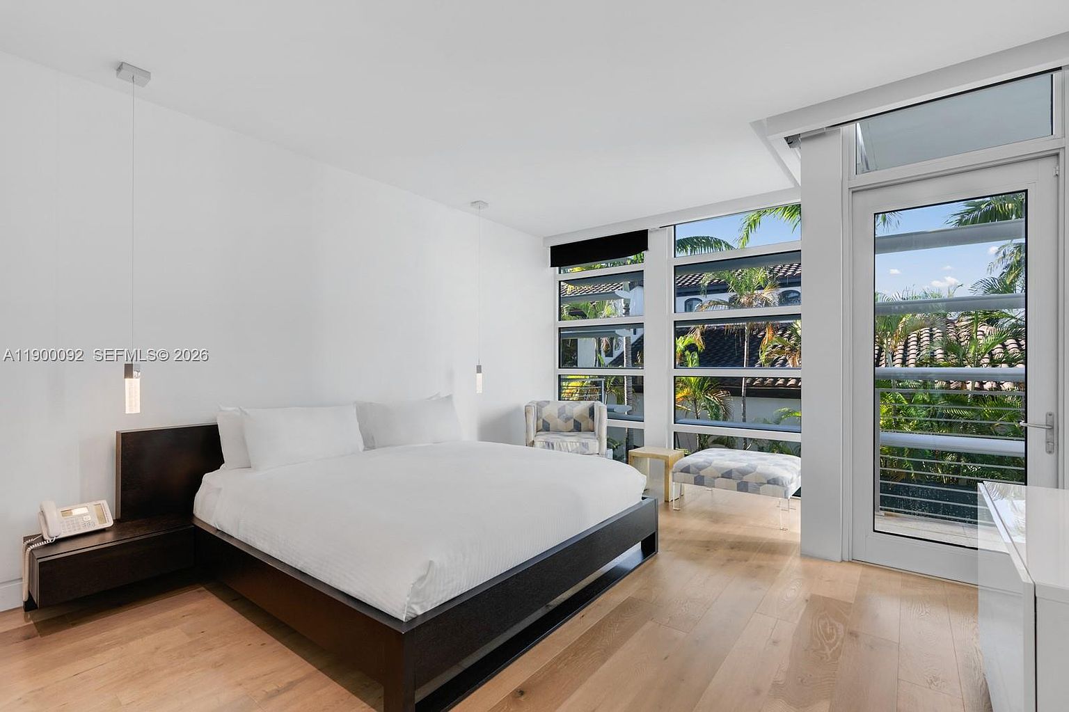 This bright and airy primary bedroom features a minimalist aesthetic with light wood flooring and crisp white walls. A dark wood bed frame provides a striking contrast to the white bedding, while large windows offer a view of lush greenery outside. The room is accented by two sleek pendant lights and a small seating area with a patterned chair and ottoman.