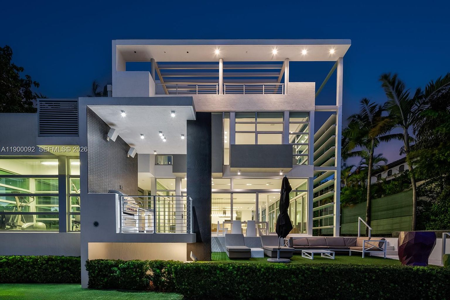 This modern, multi-level residence is captured at twilight, showcasing a sophisticated architectural design with clean lines and expansive glass walls. The rear exterior features a raised terrace with an outdoor kitchen, a lower patio area with comfortable lounge seating, and a manicured lawn bordered by lush hedges. The scene is illuminated by recessed lighting and interior glow, creating an inviting and luxurious atmosphere.