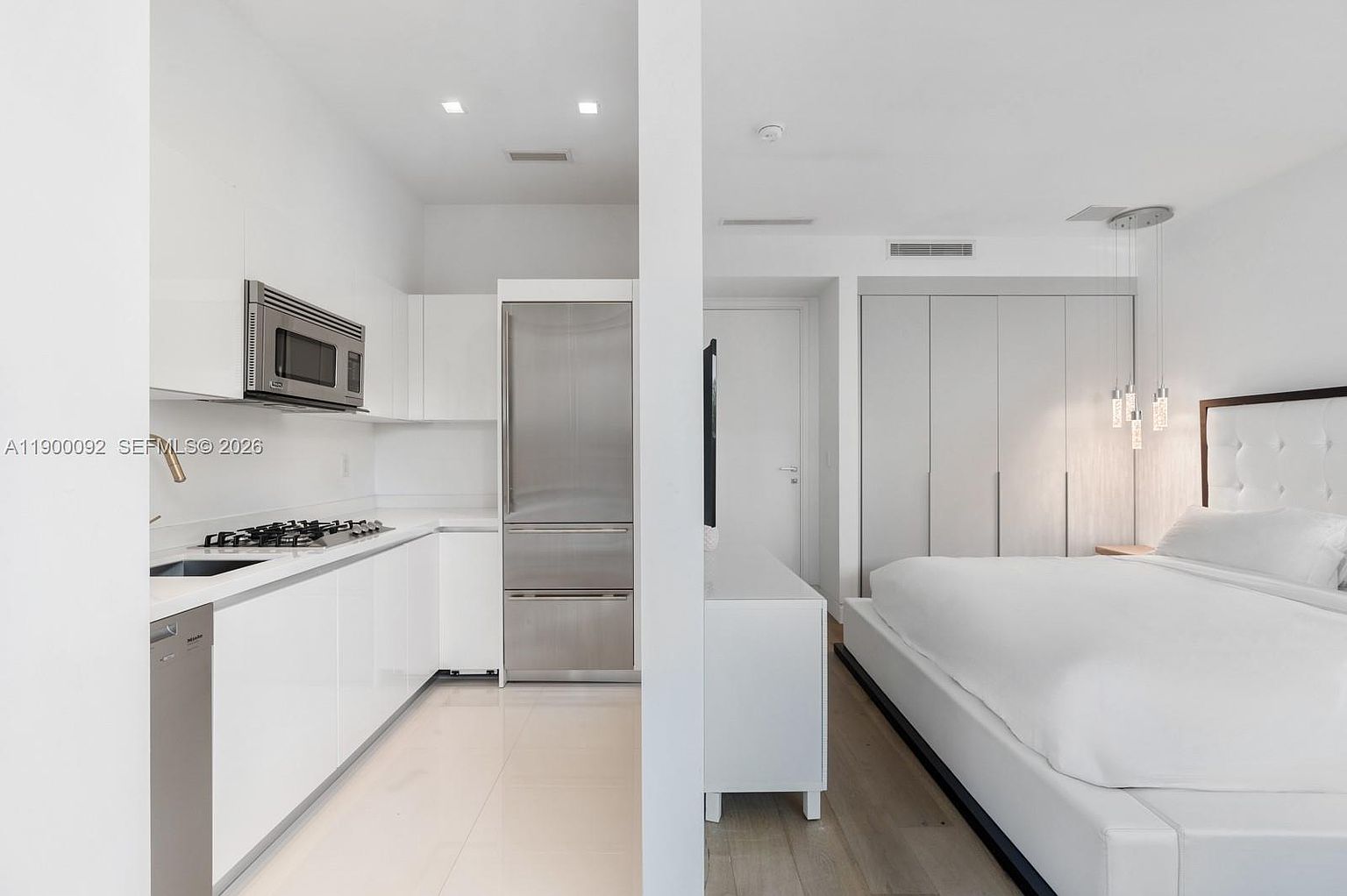 This image captures a modern, minimalist studio-style layout featuring a sleek, white-cabinet kitchen adjacent to a bedroom area. The kitchen is equipped with stainless steel appliances, including a built-in refrigerator and microwave, while the bedroom space features a plush white bed and light wood flooring. The perspective is from the kitchen looking toward the bedroom, emphasizing an open-concept design with clean lines and a bright, neutral color palette.