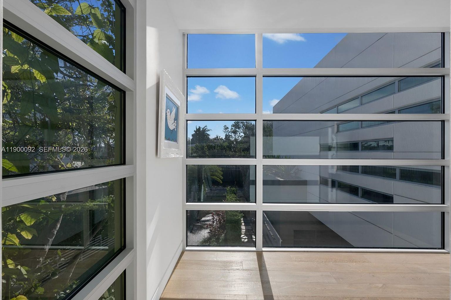 This interior space features a striking corner design with large windows that provide expansive views of the outdoors and neighboring architecture. The room is finished with light wood flooring and crisp white walls, creating a bright and airy atmosphere. A framed piece of art hangs on the wall, adding a touch of color to the minimalist, modern aesthetic.