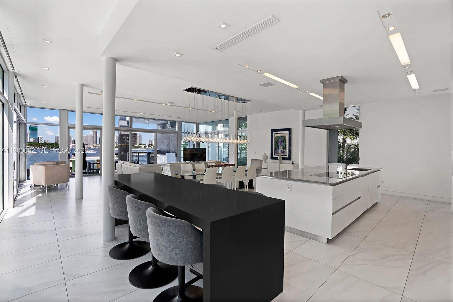 This expansive, open-concept kitchen features a sleek, black breakfast bar with grey upholstered stools, contrasting against a large white central island with a stone countertop. The space is illuminated by a modern, multi-pendant light fixture and recessed ceiling lighting, creating a bright and airy atmosphere. Large windows provide a panoramic view of the waterfront cityscape, seamlessly blending indoor luxury with the outdoor environment.