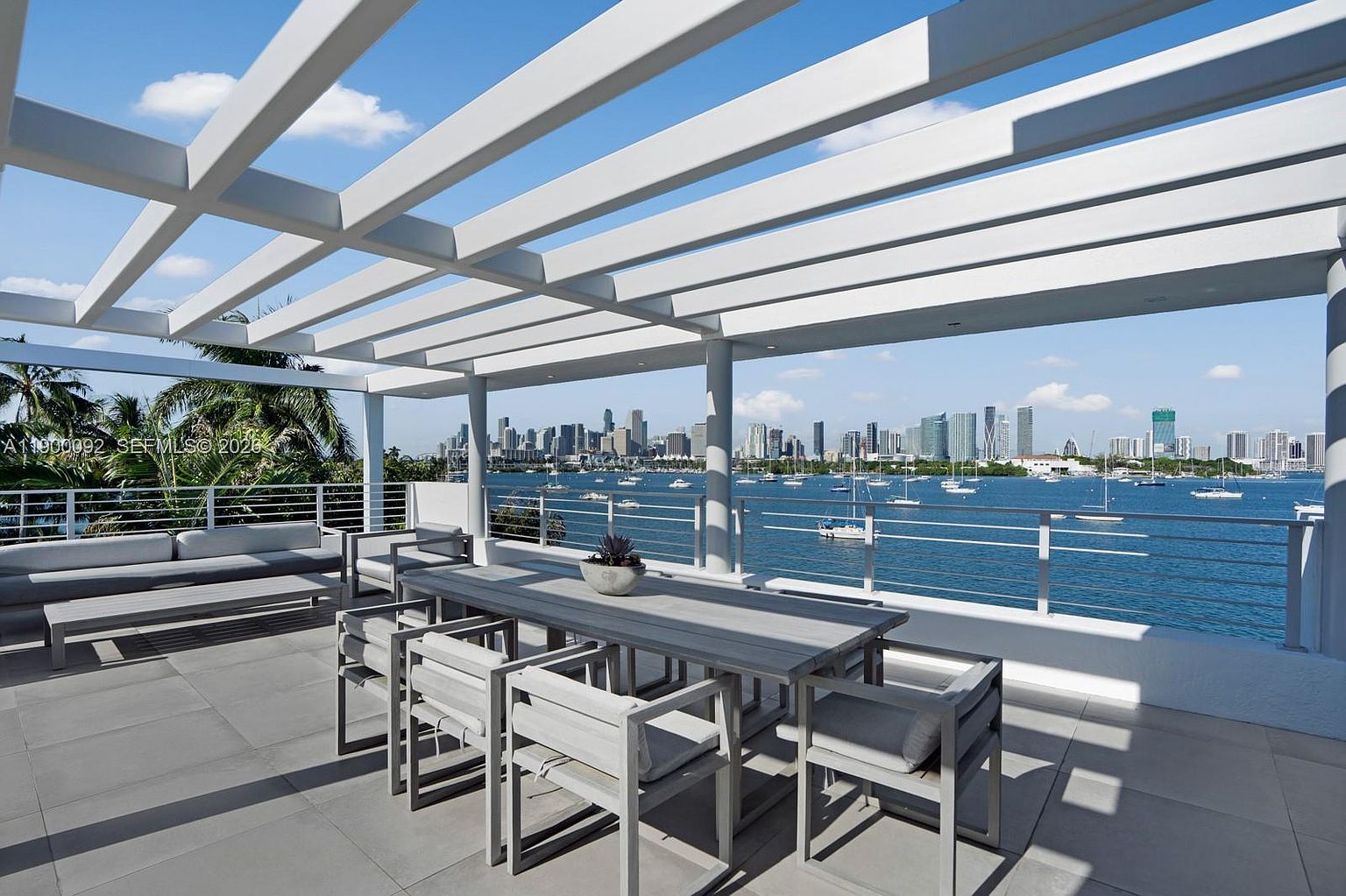 This expansive, modern balcony features a sleek pergola structure providing partial shade over a contemporary outdoor dining set and a comfortable lounge area. The space is defined by clean lines and neutral-toned tile flooring, offering an unobstructed, panoramic view of the bay and the distant city skyline. The open-air design creates a seamless transition between the living space and the waterfront environment.
