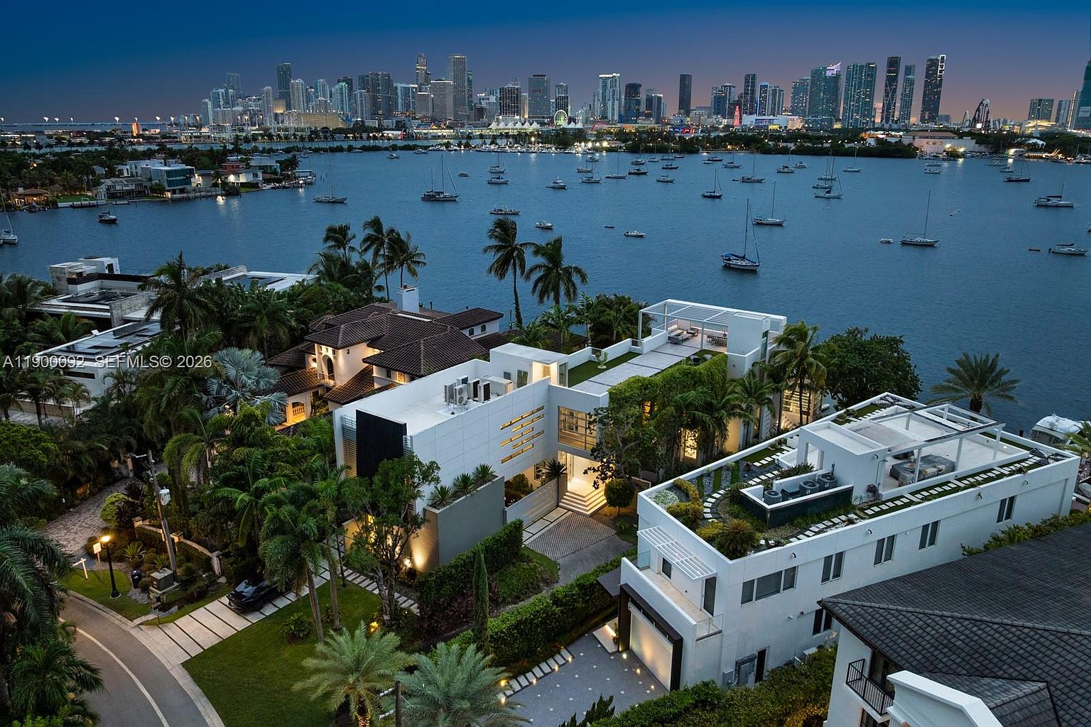 This high-angle aerial view captures a luxurious modern waterfront estate at dusk, showcasing its sleek white architecture, flat rooflines, and expansive rooftop terraces. The property is nestled among lush tropical landscaping and features a private driveway, while the background offers a stunning panoramic view of a bay filled with sailboats and the illuminated city skyline. The composition emphasizes the seamless integration of high-end residential design with its prime coastal location.
