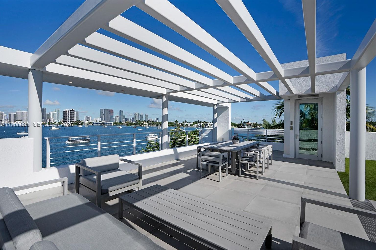 This expansive rooftop terrace features a modern pergola structure providing partial shade over a contemporary outdoor dining set and lounge area. The space is finished with large-format light-colored tiles and offers a panoramic view of the bay and distant city skyline. The perspective is captured from a slightly elevated eye-level, emphasizing the seamless transition between the private outdoor living area and the scenic waterfront backdrop.