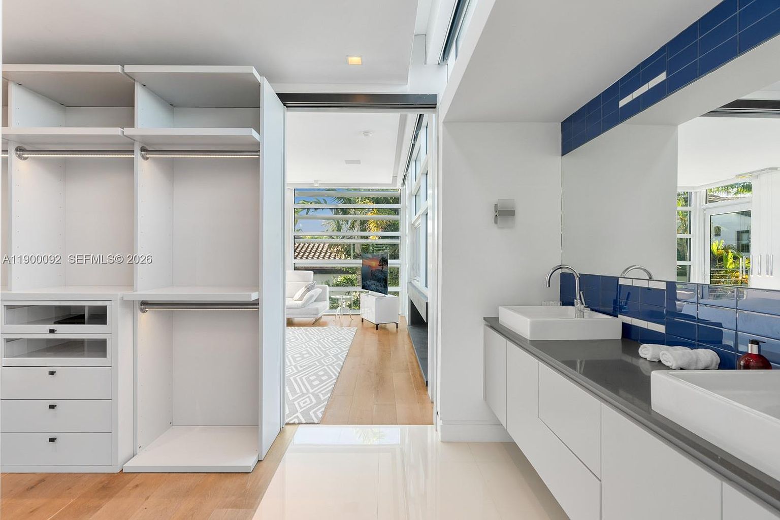 This modern bathroom features a dual-sink vanity with white cabinetry and a dark stone countertop, accented by a striking blue tiled backsplash. To the left, a spacious, open-concept walk-in closet with built-in shelving provides seamless storage. The perspective looks through a wide doorway into an adjacent living area, which is bathed in natural light from large windows overlooking a lush outdoor setting.