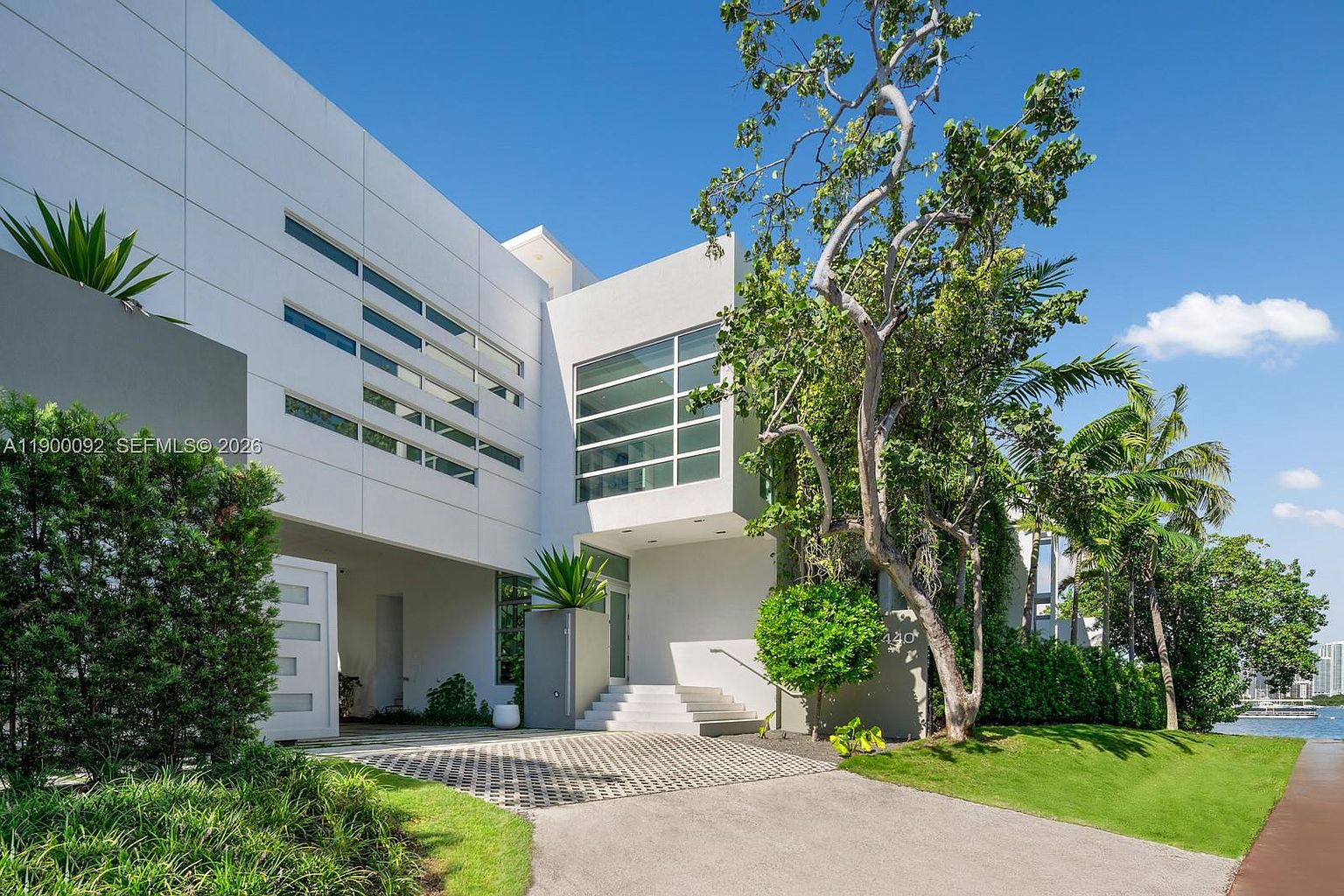This modern, white-stucco residence features a striking geometric design with clean lines and large, multi-paned windows. The property includes a paved driveway leading to a covered entryway with steps, surrounded by lush tropical landscaping and mature trees. The perspective captures the home from a low angle, emphasizing its architectural scale and proximity to the waterfront.
