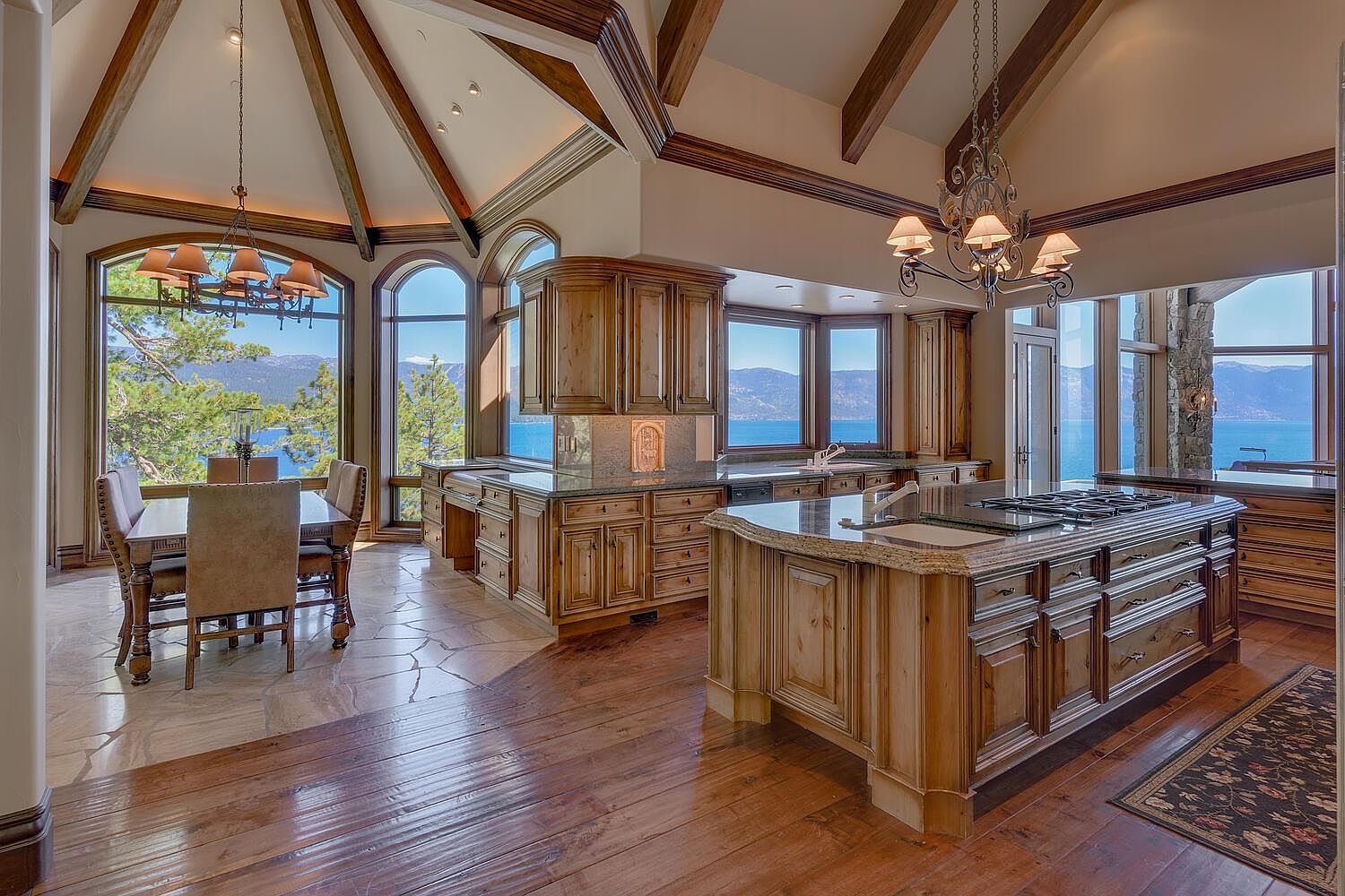 This is a spacious, luxury kitchen with custom wood cabinetry, granite countertops, and high-end appliances. The kitchen features a large island with a cooktop, a dining area with large windows offering stunning lake views, and exposed wooden beams on the vaulted ceiling. The overall impression is one of elegance and sophistication, perfect for entertaining and enjoying the scenic surroundings.