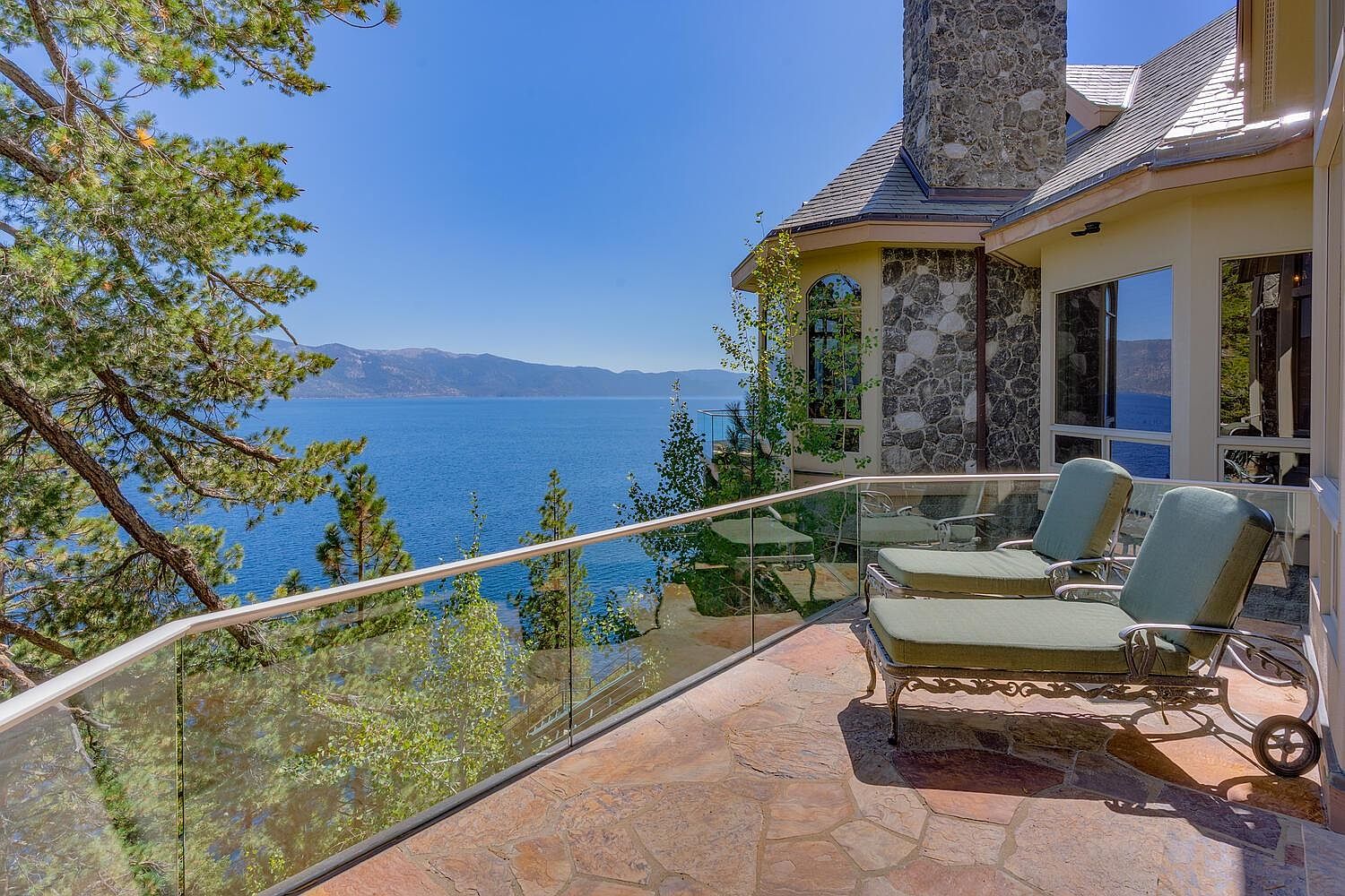 This image showcases a luxurious patio or balcony with stunning lake views. The space features comfortable lounge chairs, a glass railing, and a stone tile floor, creating an inviting outdoor living area. The architectural details of the house, including stone accents and large windows, add to the overall elegance and appeal of the property.