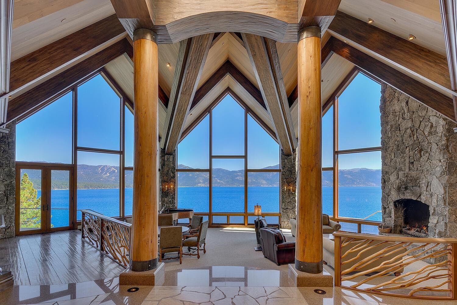 This grand living room boasts floor-to-ceiling windows offering stunning lake views, complemented by exposed wooden beams and stone accents. The room features a mix of seating arrangements, including chairs and sofas, arranged around a central area. The polished stone flooring and natural wood railings add to the luxurious and rustic ambiance, creating a warm and inviting space.