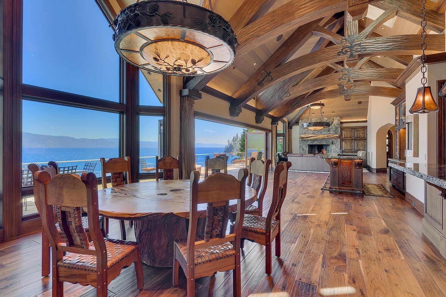 This is an interior shot of a dining room featuring a large, round wooden table with unique chairs. The room boasts expansive windows offering a stunning view of the water and sky. The ceiling has exposed wooden beams and ornate light fixtures, adding to the room's rustic yet luxurious ambiance.