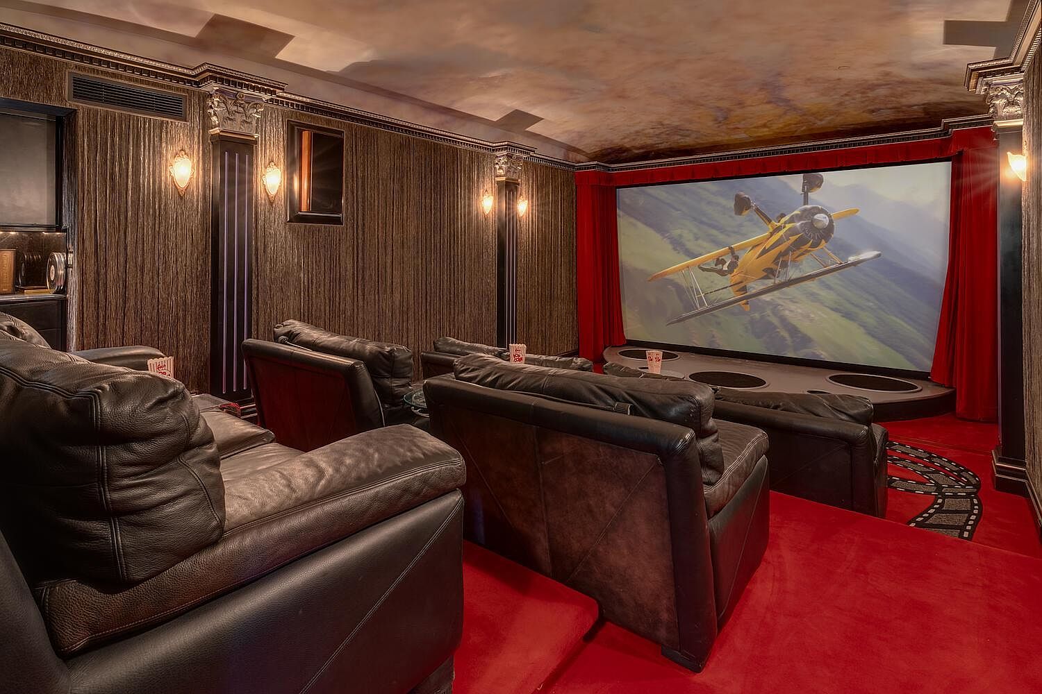 This is an interior shot of a home theater. The room features plush leather seating arranged in rows facing a large screen displaying an image of a yellow airplane. The walls are dark and textured, with decorative columns and sconces providing ambient lighting, and the floor is covered in red carpet, creating a luxurious and immersive viewing experience.