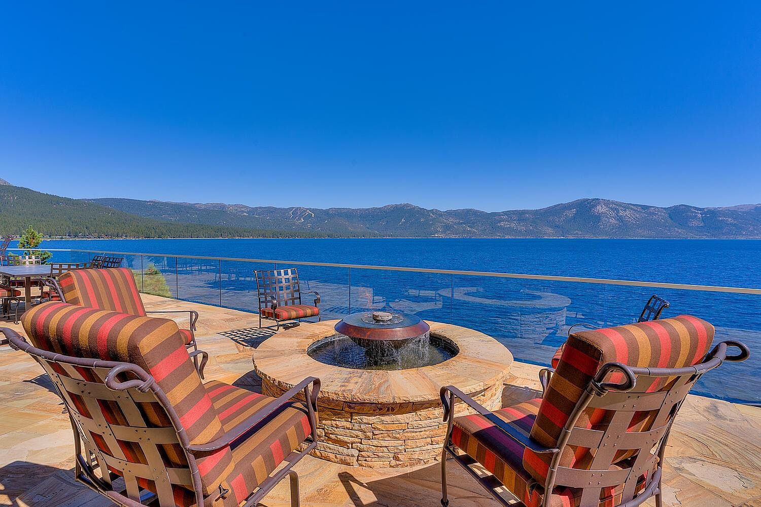 This image showcases a luxurious outdoor patio or deck area with stunning lake views. The focal point is a stone fire pit with a water feature, surrounded by comfortable striped armchairs. A glass railing provides an unobstructed view of the lake and mountains in the background, creating a serene and inviting atmosphere.