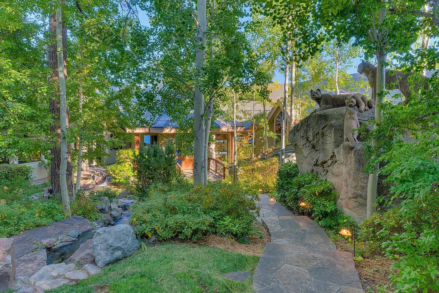 This exterior shot showcases a beautifully landscaped yard and garden, featuring a stone pathway leading towards a charming home. A small stream with rocks adds a natural element, while mature trees and lush greenery create a serene and private atmosphere. A unique rock formation with bear statues adds a whimsical touch to the property.