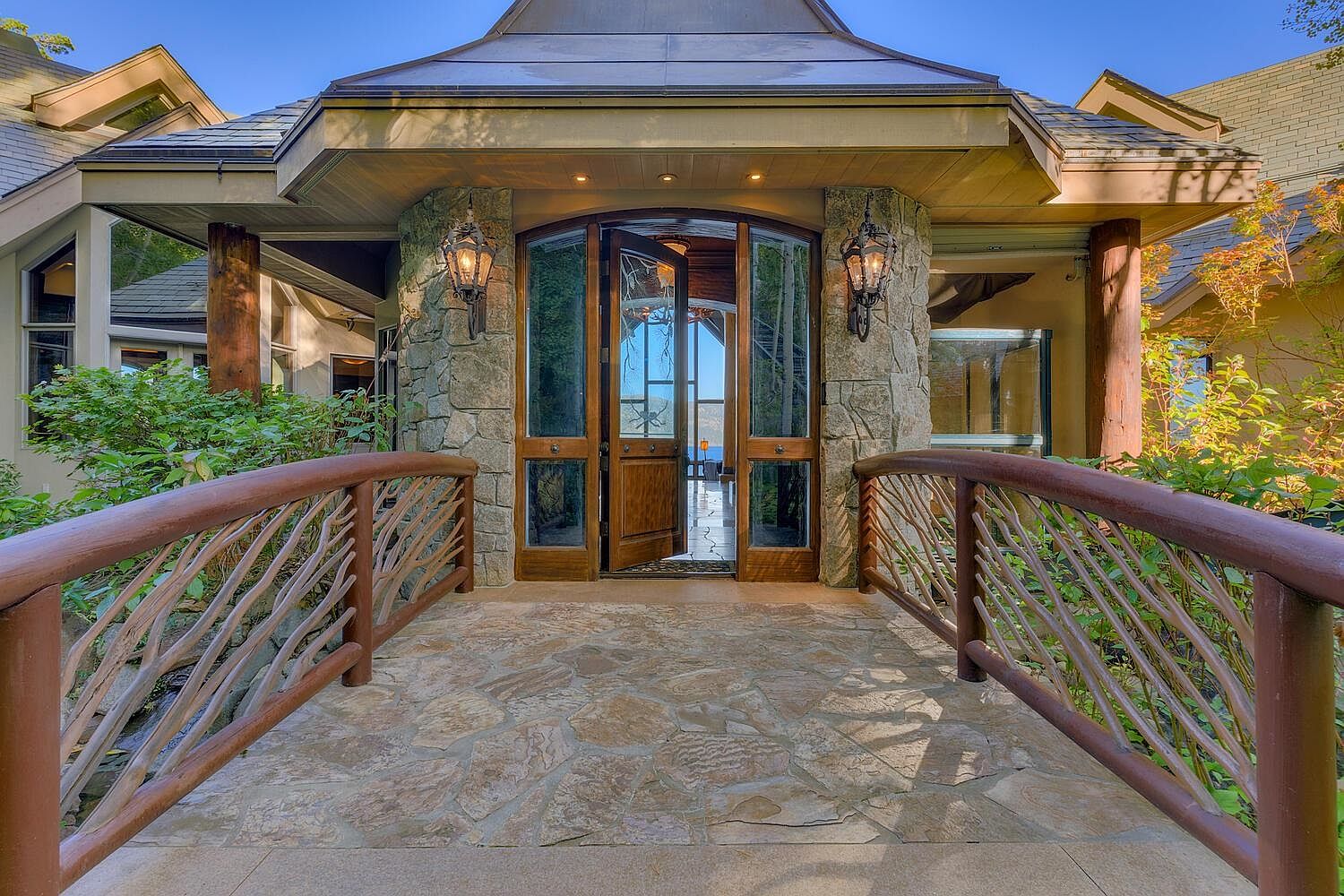 This image showcases the grand entryway of a luxurious home. A stone pathway leads to an arched wooden door framed by stone pillars and elegant lanterns. The unique wooden railing adds a rustic touch, while the overall design exudes sophistication and curb appeal.