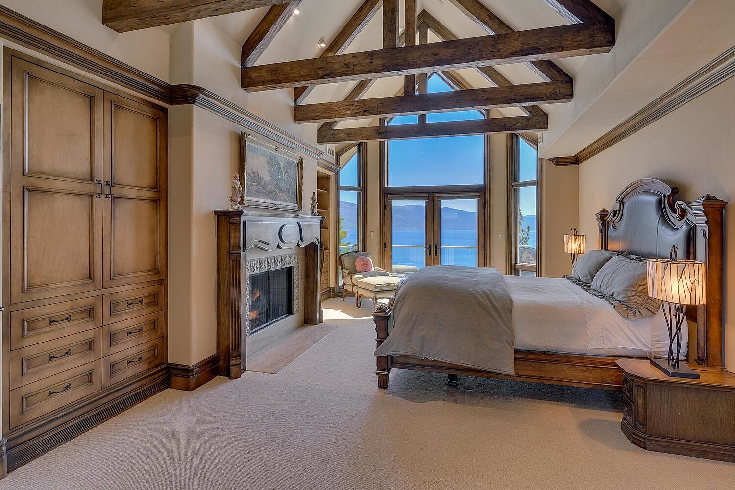 This is a luxurious primary bedroom featuring a king-size bed with a dark wood frame and a tufted headboard. The room has a fireplace with an ornate mantel, built-in cabinetry, and large windows offering a view of the water. Exposed wooden beams add a rustic touch to the high ceiling, creating a warm and inviting atmosphere.