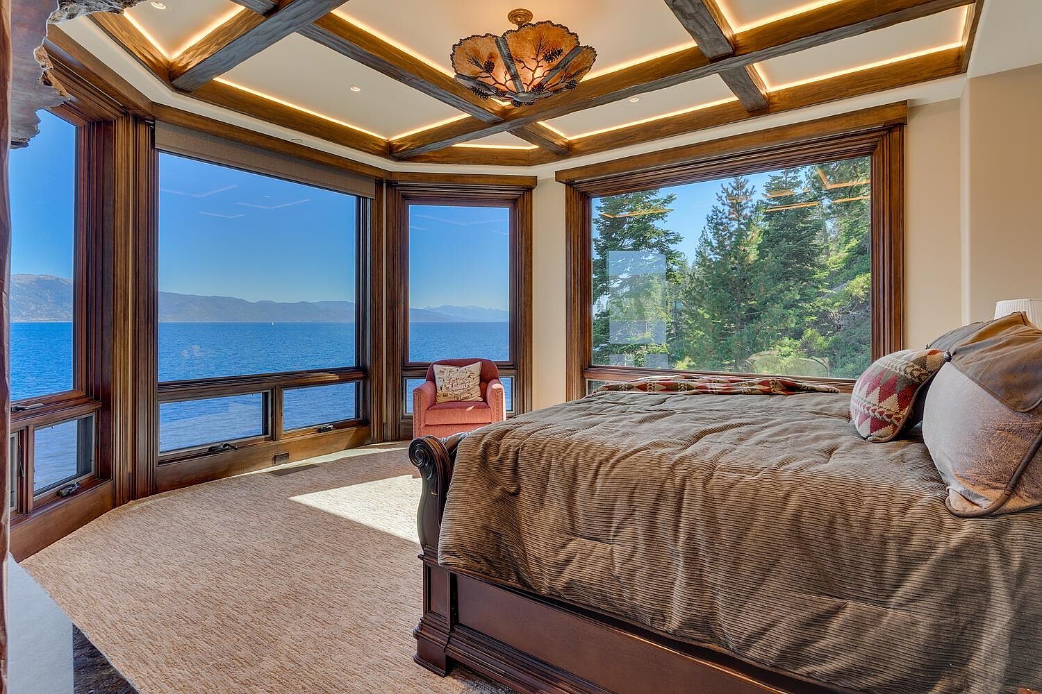 This is a luxurious primary bedroom featuring a large bed with a brown comforter and decorative pillows. The room boasts expansive windows offering stunning views of a lake and mountains, complemented by a coffered ceiling with ambient lighting and a unique chandelier. The overall impression is one of comfort, elegance, and breathtaking scenery.
