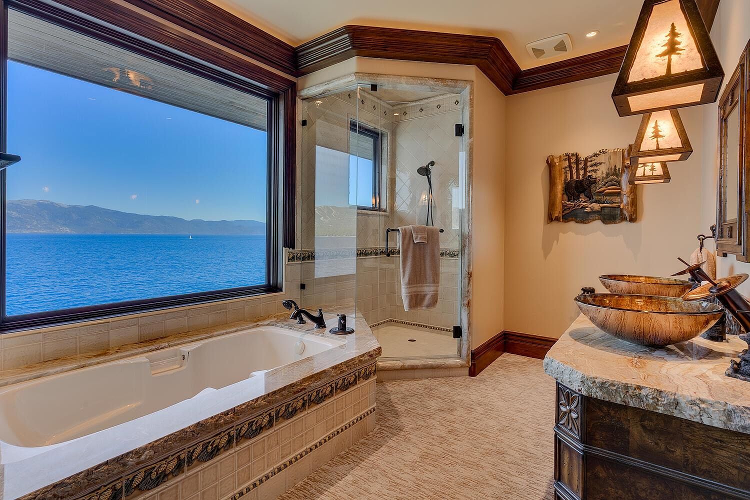 This is a luxurious primary bathroom featuring a large soaking tub positioned beneath a wide window with a scenic water view. A glass-enclosed shower is adjacent to the tub, and a double vanity with unique vessel sinks and decorative lighting completes the space. The bathroom exudes a spa-like atmosphere with its warm color palette and high-end finishes.