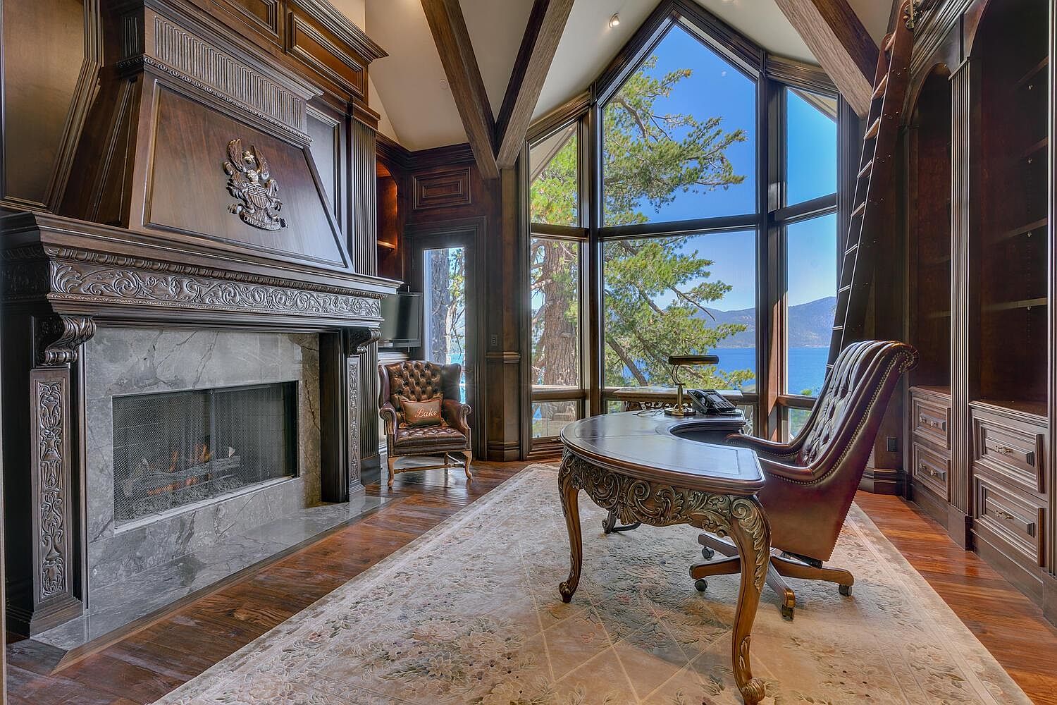 This is a luxurious home office featuring a large wooden desk and leather chair, complemented by a grand fireplace and built-in bookshelves with a ladder. The room is bathed in natural light from a large window offering a scenic view of a lake, creating a sophisticated and inviting workspace. The overall impression is one of elegance and productivity.
