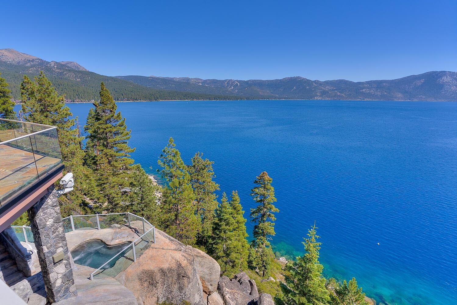 This image showcases a stunning patio or deck area with a built-in spa overlooking a vast, deep blue lake and mountain range. The patio features stone elements and glass railings, providing unobstructed views. The overall impression is one of luxury and tranquility, highlighting the property's prime location and outdoor living potential.